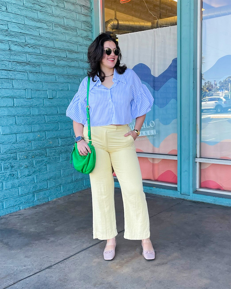 Woman wearing striped light blue blouse, yellow pants, green handbag and pink ballet flats 