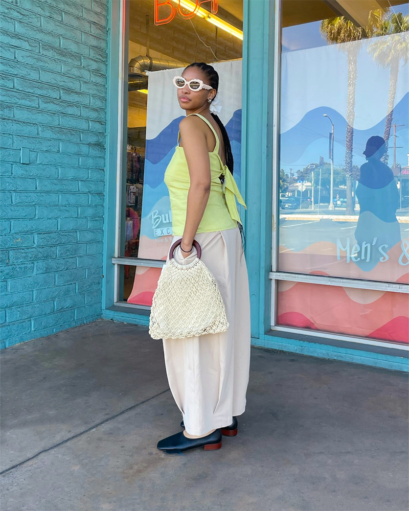 A woman wears a light green sleeveless top, beige pants, a woven tote bag and black flat shoes