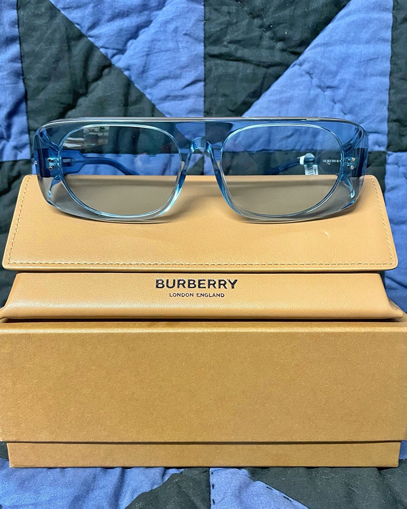 Blue tinted Burberry sunglasses sit on top of their box with a quilt in the background