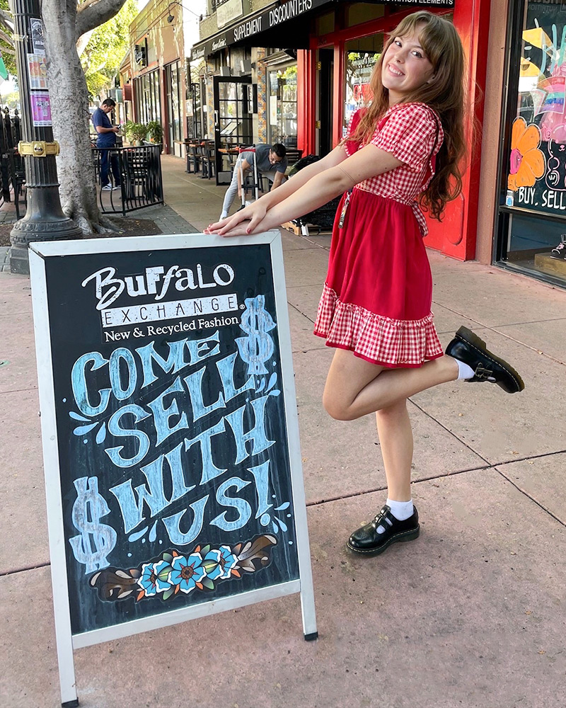 A woman poses with a sidewalk sign that reads "Buffalo Exchange Come Sell With Us $" 