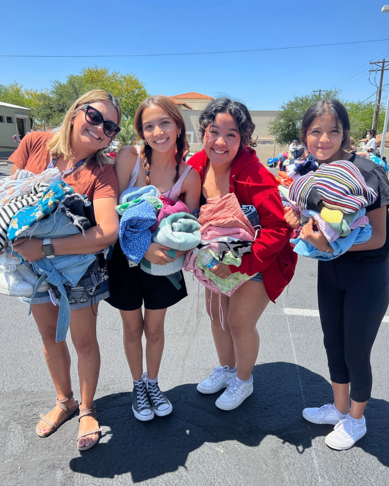 a group of friends standing in parking lot smile while holding piles of clothing 