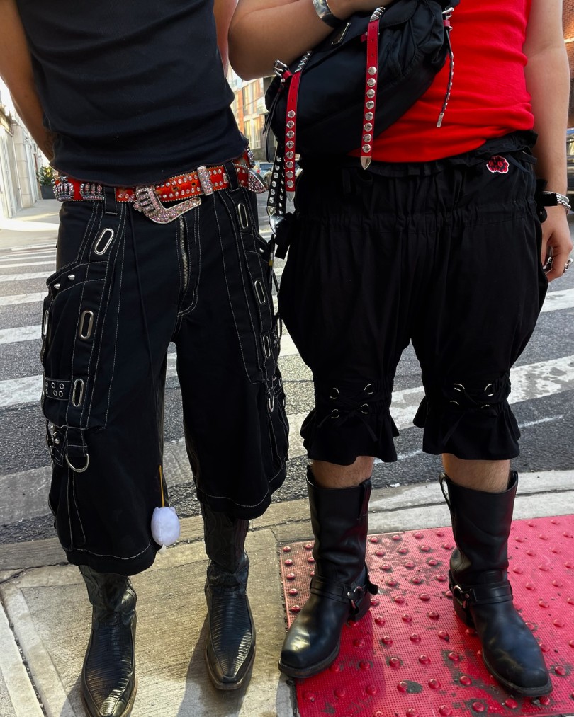 A close up of two people dressed in red & black moto style on a New York City street corner wearing tanks, shorts with grommet details, and leather harness boots