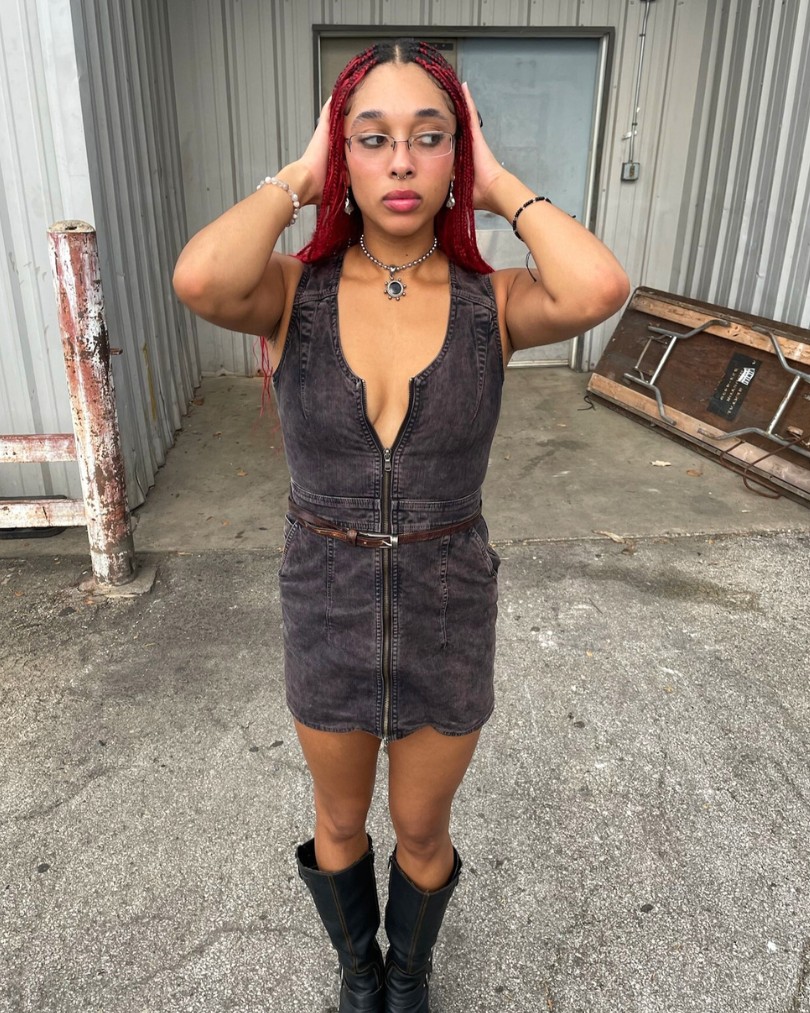 Person standing in front of an industrial building while wearing metal framed glasses, an acid wash denim dress, leather knee-high moto boots and silver jewelry