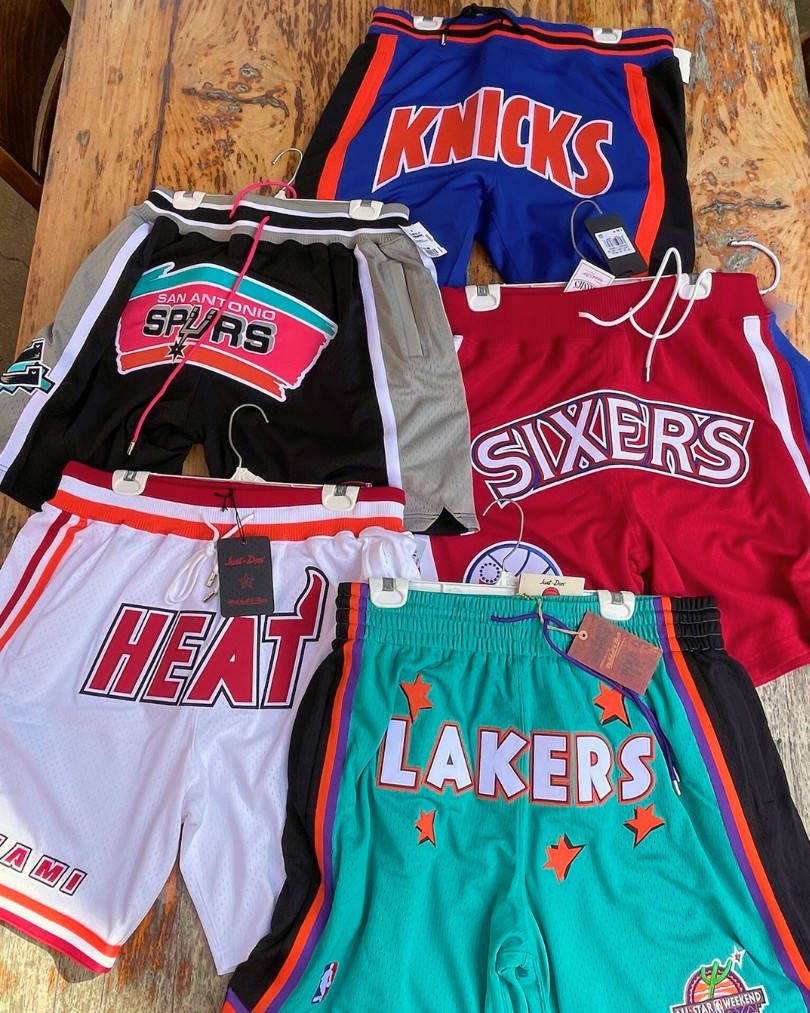 A collection of NBA-branded basketball shorts for teams like the New York Knicks, Miami Heat, Los Angeles Lakers, Philadelphia 76ers and San Antonio Spurs from Mitchell & Ness laid flat on a wooden table