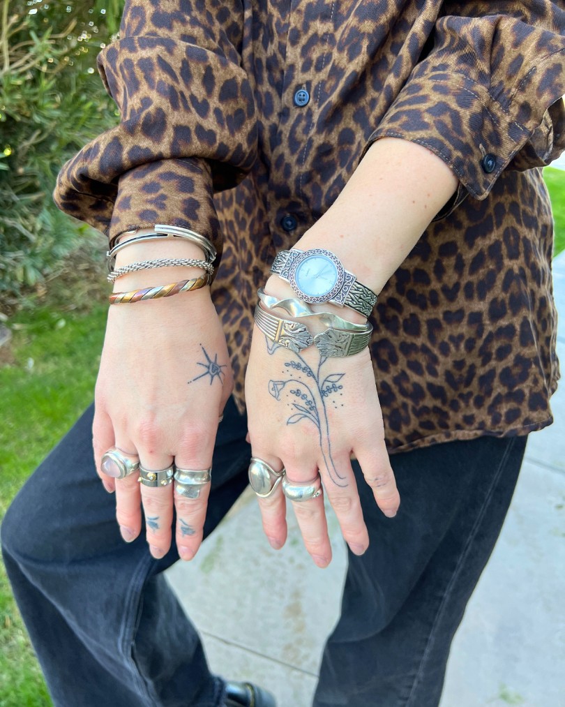 Close up of a person's hands with stick and poke tattoos & mixed metal jewelry including sterling silver bracelets, a vintage pearl-faced watch and chunky silver rings